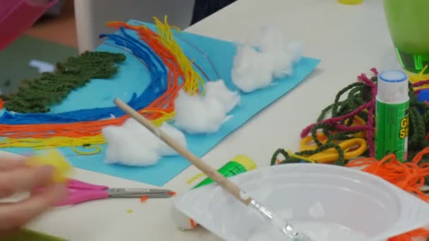 Kid 's Hands Put a Rainbow-Applique to a Table Applique of Colorful Threads Cotton Clouds Hanks of Threads Are on a Table Bowl of Glue and Brush Classroom — стоковое видео