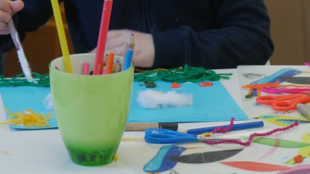 Руки ребенка Положите клей с кистью на чашку голубой бумаги карандаши Дети делают радугу-аппликацию разноцветные нити ножницы нити на столе — стоковое видео