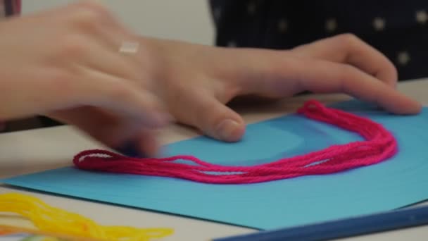 Las manos de las niñas están pegando un hilo amarillo a los rosados haciendo un arco iris-apliques en una hoja azul de papel poniendo un pegamento que la gente está haciendo regalos hechos a mano — Vídeo de stock