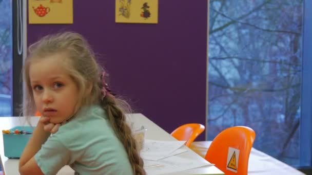 Niño se ha inclinado a sus brazos y mirando hacia atrás Niño está sentado en la mesa en el aula Lápices coloridos en una caja Chica rubia está dibujando en la biblioteca — Vídeos de Stock