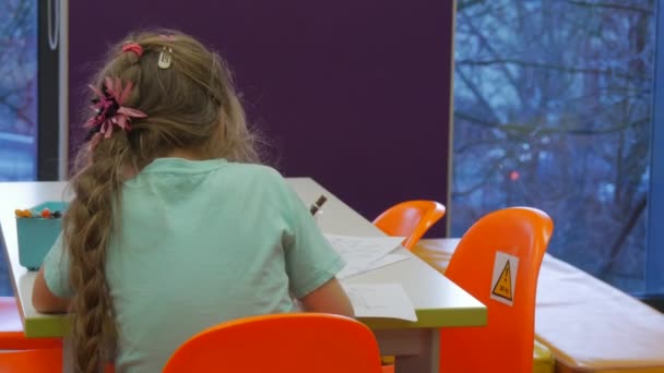 Niño está dibujando con su espalda a la cámara Lápices de colores Papel en una mesa Niño está sentado en la mesa en el aula Chica rubia está dibujando en la biblioteca — Vídeos de Stock