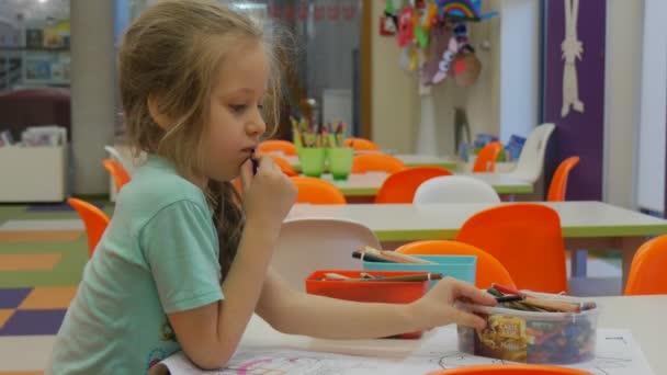 Малюк посміхається накладає олівець на коробку, що грає з олівцями, сидячи за столом у класі маленька дівчинка блондинка грає в бібліотеці — стокове відео