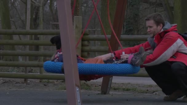 Kid is Sitting on a Swing Father is Push Her Daughter is Playing on a Swing in a Park Child and Dad Are Laughing Family Have a Rest on a Playground — Vídeo de Stock