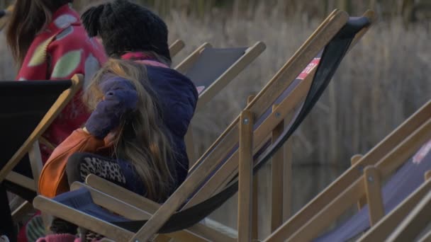 Chica está sentada en silla y retumba en la bolsa La gente está sentada alrededor de su niño rubio Descansando en el muelle Hora de la noche Día soleado Opole Polonia al aire libre — Vídeos de Stock