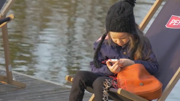 Enfant est assis dans la chaise longue et jouer au téléphone fille blonde reposant sur la jetée eau calme sur un fond Soirée Heure Ensoleillé Jour Opole Pologne En plein air — Video