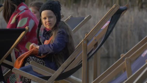 Dítě sedí v křesle a prohrabává v sáčku lidé sedí kolem její blond dívka spočívají na Pier večer čas slunečný den Opole Polsko venku — Stock video