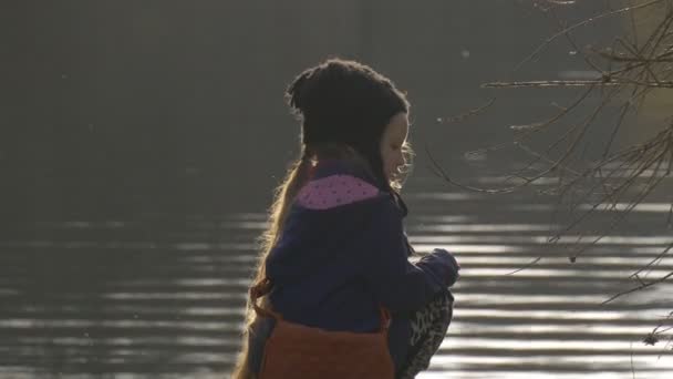 Kleines Mädchen sitzt aufrecht auf einem grünen Ufer eines Sees zu Fuß in der Nähe von ruhigen ruhigen friedlichen Teich plätschert auf dem Wasser Abend Sonnenuntergang Frühling — Stockvideo