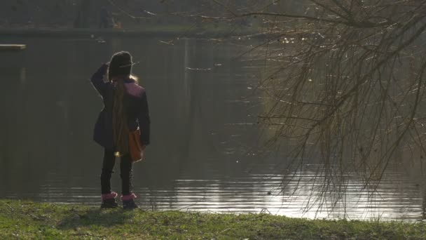 Kind steht in der Nähe des Sees spazieren gehen in der Nähe der ruhigen ruhigen friedlichen Teich plätschert auf dem Wasser grünes Gras nackte Äste Abend Sonnenuntergang Frühling — Stockvideo