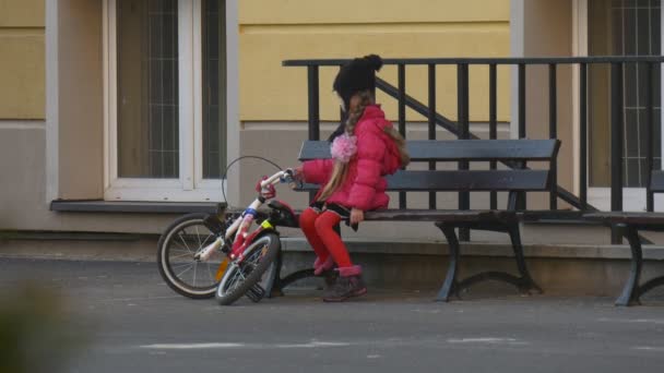 Kid flicka sitter på en bänk röra en cykel har en vila efter ridning en cykel i kväll asfalterade vägen ungen är att lära sig cykla — Stockvideo