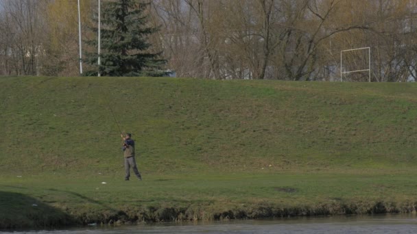 Man slingerbewegingen met Fishing Rod gooit aas permanent op tegenovergestelde oever van een rivier op een groene weide Fir-Tree groeit op een heuvel vlaggenmast met zonnige dag van de vlag zwaaien — Stockvideo