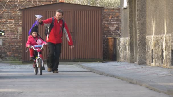 Far är kör längs dotter rider på cykel lär hans dotter att rida en cykel Kite är fast på cykel barn rider på en cykel Sunny Day fjäder — Stockvideo