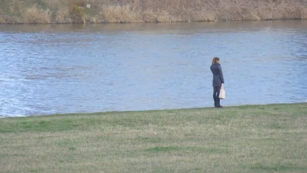 女人站在河银行斗牛方法女人人是走与狗的绿色草地上在河银行水流动温和的冬天 — 图库视频影像