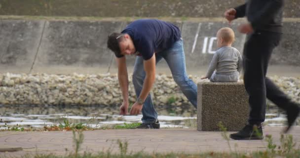 Little Boy está sentado em pedra quadrada na margem do rio e joga as pedras que o homem dá algumas pedras para o menino As pessoas estão andando andando de bicicleta — Vídeo de Stock