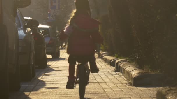 Holčička je na kole dál podél ulice v Opole Polsko dítě je na koni podél the automobily zaparkované na ulici silueta sluneční paprsky sluneční den jaře — Stock video