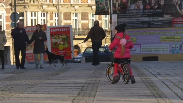 Niña está montando la bicicleta Día de la ciudad en Opole Polonia Los peatones pasean a los perros Niño está montando entre la multitud de personas que caminan por una calle Día soleado — Vídeos de Stock