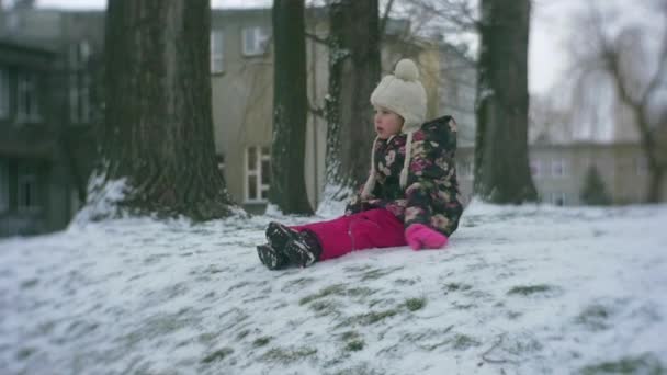 Kleines Mädchen sitzt auf einem Schnee auf einem Hügel im Freien Winter Zeitlupe dreht sich um Gebäude Schnee frostiges Mädchen in geblümter Jacke rosa Hose — Stockvideo