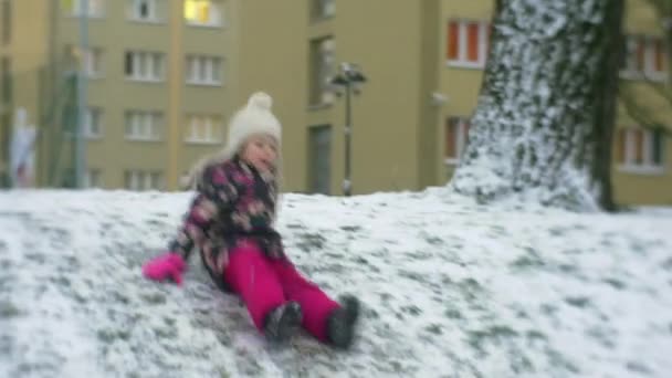 Happy Little Girl corre diapositivas cuesta abajo por el hielo levantándose y caminando Smiling Buildings invierno Nieve Frosty Girl en chaqueta de flores y pantalones rosados — Vídeo de stock