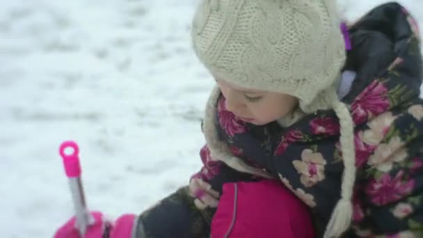 孩子挖雪与粉红色铲是一个女孩玩户外坐在她膝盖冬建筑物雪霜姑娘在花外套白色帽子 — 图库视频影像