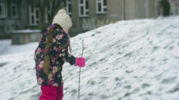 Küçük kız Hill ile bir şube sopa yürüyerek kar sürünen kış binalar kar buz gibi kız çiçekli ceket ve pembe pantolon kadar tırmanışı mı — Stok video