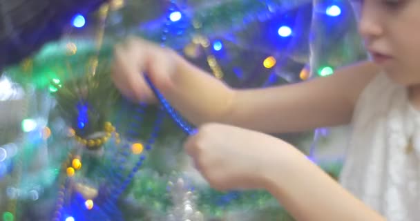 Ξανθό κορίτσι λύνει ένα μπλε χάντρες γιρλάντα θέτοντάς την σε μια για το νέο έτος δέντρο πεύκων χρυσή καμπάνα-γιρλάντα και πολύχρωμο Lamplights οικογένεια Χριστουγεννιάτικη γιορτή — Αρχείο Βίντεο