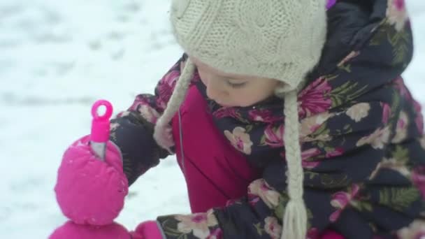 Niña está cavando una nieve con una pala rosa Kid está jugando al aire libre sentada en sus rodillas Edificios de invierno Nieve Frosty Girl en chaqueta floreada Sombrero blanco — Vídeo de stock
