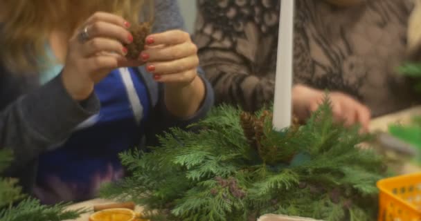 Erwachsene Frau wählt Tannenzapfen und flechtet Draht drinnen, legt ihn zur weihnachtlichen Komposition von Adventskranzzweigen auf den Tisch des Neujahrsmeisterkurses — Stockvideo