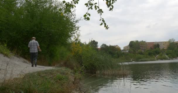 L'anziano uomo anziano sta camminando lungo il sentiero lungo il fiume Lago Ondeggiante acqua Case rosse Edifici presso la riva opposta sulla collina verdi freschi cespugli — Video Stock