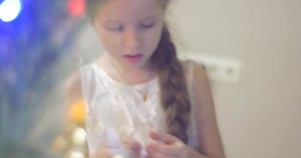 Girl is Putting a Golden Bells-Garland Ramas de pino de árbol de año nuevo en la celebración familiar en casa Pequeña chica rubia curiosa en vestido blanco — Vídeo de stock