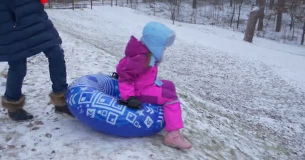 Malá holčička jezdí z kopce na nafukovací sněžnou Sáňku skoro zaklepá na muže, který stojí na úpatí kopce zasněžený zimní den — Stock video