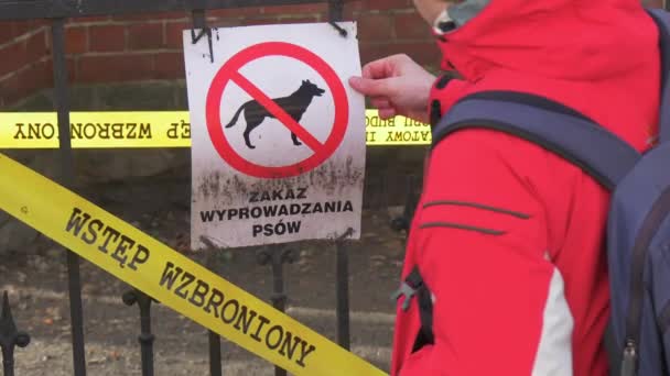 Mann streift Warnschild Kein Hundeschild mit gelbem Zaunband Aufschrift in polnischer Sprache Backpacker in roter Sportjacke kommt und läuft davon — Stockvideo