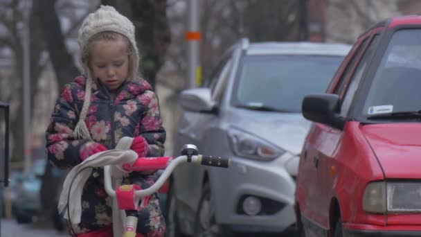 Bambino Unties suo sciarpa andando a cavalcare un bicicletta città strada automobili sono parcheggiati lungo un marciapiede cartelli paesaggio urbano bambina in giacca fiorita sta giocando — Video Stock