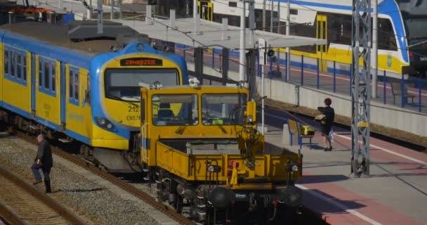 Mujer en Platphorm mira el reloj El hombre cruza el ferrocarril Dos trenes eléctricos de pasajeros azules y amarillos están de pie en la locomotora de la estación de tren — Vídeo de stock