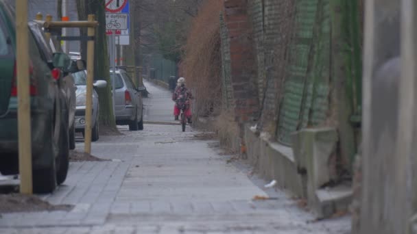 El niño está montando lejos de una bicicleta y detiene a los coches de la calle de la ciudad están estacionados a lo largo de una acera Señales de tráfico Paisaje urbano Niña con chaqueta floreada está jugando al aire libre — Vídeos de Stock