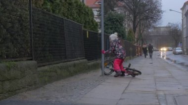 Düşen çocuk kalkar yürüyüş ağlamaya oldu At Binme Bisiklet şehir sokak arabalar vardır park boyunca bir kaldırım, küçük kız çiçekli ceket tarafından oynarken açık havada olduğunu