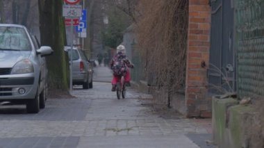 Çocuk sürme bir bisiklet uzak şehir sokak yol işaretleri arabalar vardır park boyunca bir kaldırım kırmızı tuğla çit Cityscape, küçük kız çiçekli ceket tarafından oyun olduğunu