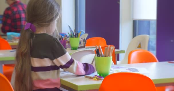 Menina senta-se em uma cadeira laranja perto da mesa branca — Vídeo de Stock