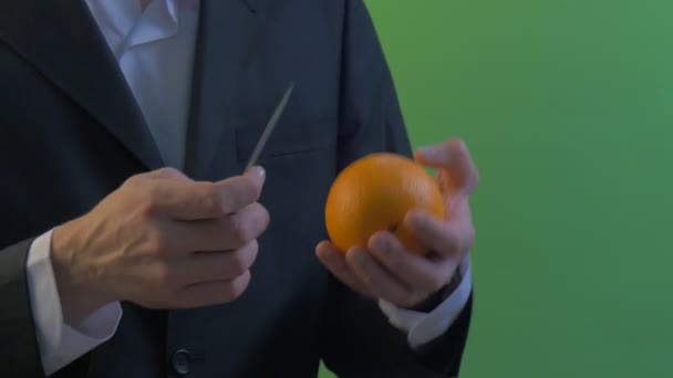 L'homme vient s'asseoir et éplucher une orange avec un couteau sur un écran vert Jeune homme d'affaires en chemise blanche et costume noir main en anneau d'or sur un doigt — Video