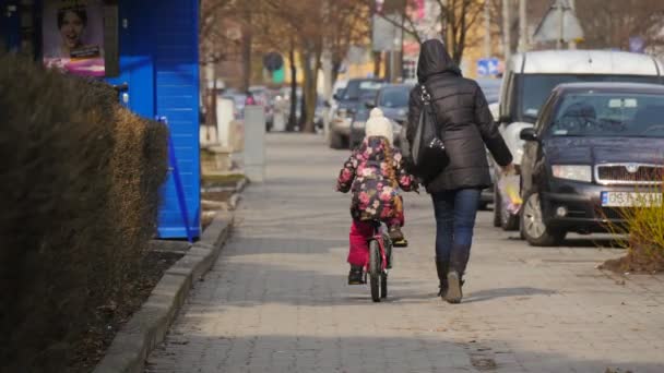 Люди Мама и дочь на городской улице пешеходы припаркованные автомобили городской пейзаж Маленькая девочка верхом на велосипеде женщина идет вдоль ребенка на обочине — стоковое видео