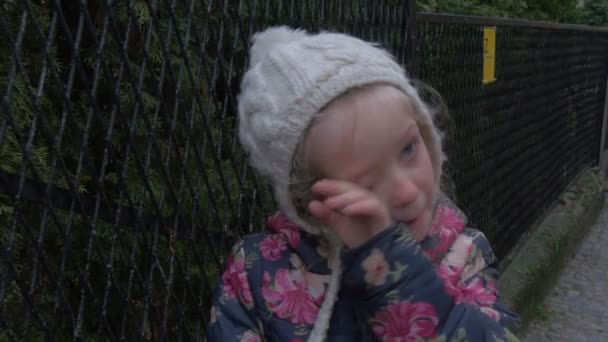 Triste enfant pleure essuie les larmes petit enfant joue en plein air blonde enfant en veste fleurie et blanc chapeau rouge visage ennuyé enfant debout à la clôture — Video