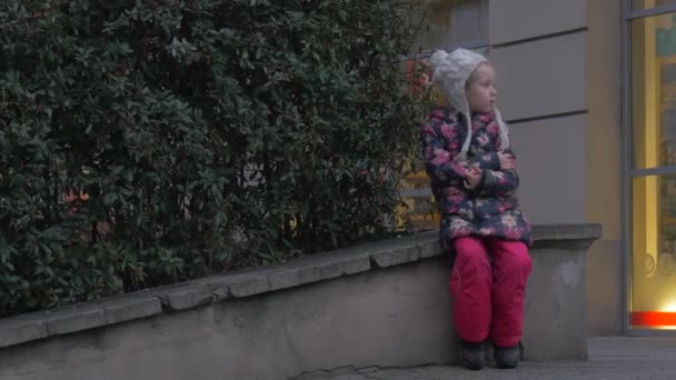 Miúda entediada está sentada em um parapeito criança brinca ao ar livre loira criança em casaco florido e chapéu branco está sentado com os braços cruzados inverno — Vídeo de Stock