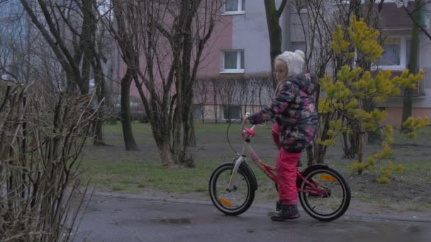 Kind fährt Fahrrad durch Park Allee Bürgersteig kahle Äste Bäume und Büsche Gebäude hinter den Bäumen Stadtbild kleines Mädchen in geblümter Jacke Abenddämmerung — Stockvideo