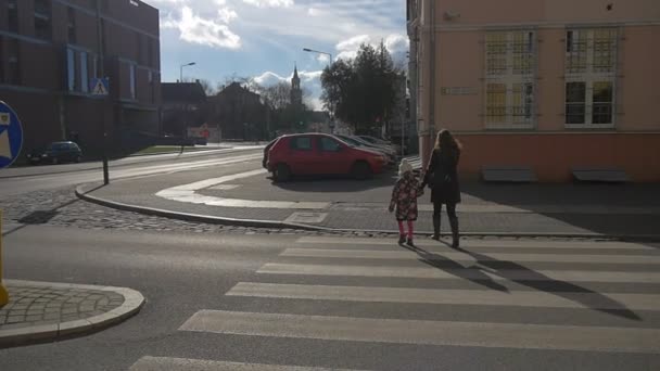Mãe e filha cruzar a estrada vazio cidade rua em Opole Bell Tower em um horizonte pedestres calçada estacionado carros nu ramos árvores dia ensolarado inverno — Vídeo de Stock