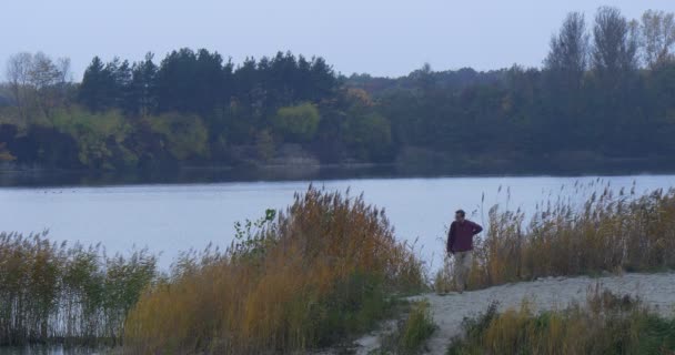 Człowiek stoi na chwilę w pobliżu brzegu rzeki w Sandy jezioro krajobraz turystycznych spacerów od drzewa sylwetki Zmierzch zielone i żółte trawa trzciny gładkiej wodzie jesieni — Wideo stockowe