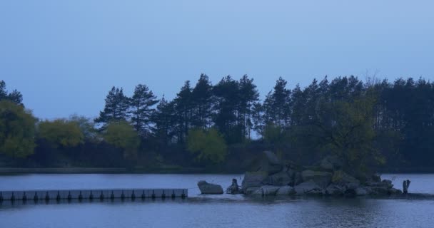 Muelle en medio de un río Pequeña isla pedregosa cubierta de árboles Paisaje otoñal Hojas amarillas y verdes Árboles al atardecer en el lado opuesto Cielo azul — Vídeos de Stock