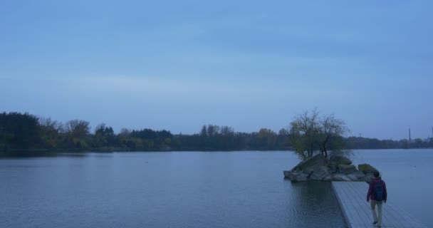 夕方に向かい側の地平線の男にコンクリート桟橋ストーニー小さな生い茂った島の木の秋の風景黄色と緑の木の上を観光客が歩きます — ストック動画