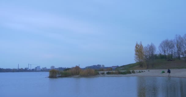 Turysta przychodzi do Sandy Bank porośnięty drzewami krzewy jesień krajobraz żółty i zielone liście Cityscape budynki sylwetki na wieczór Horizon — Wideo stockowe