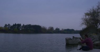 Young Man Reads a Book on a Pier at the River Stony Island Bare Branches Trees Reader at the Autumn Landscape Yellow and Green Trees Ona Horizon Dusk