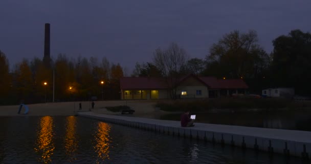 Man Works on Laptop on Concrete Pier Freelancer Works at Boat Station River Evening People Siluetas Luces de paisaje de otoño en el edificio Sandy Beach — Vídeo de stock