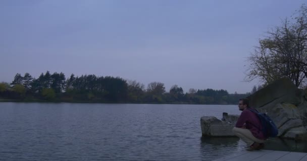 Le touriste se trouve sur la jetée de béton près de la rivière quitte la jetée Branches nues Arbres sur une petite île pierreuse Paysage d'automne Soir Arbres sur le côté opposé — Video