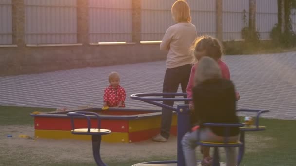 子供たちは庭の遊び場で遊んでいる、遊んで、カルーセルでスイング、ピンクのブラウスの女の子はカルーセルを動かしている、黒の女の子は座っている — ストック動画